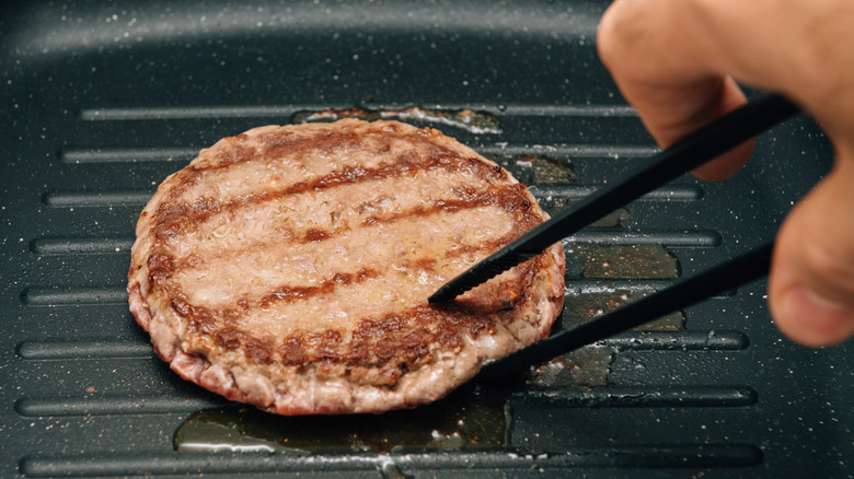 A person using tongs to flip a burger in a cast iron skillet
