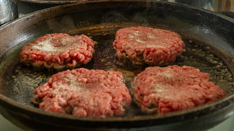 Four seasoned burger patties with dimples cooking in a cast iron skillet