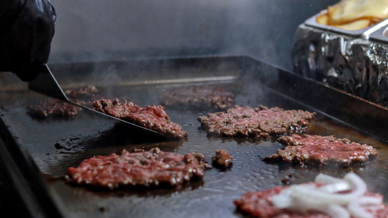 A person using a metal spatula to flip burgers on a cast iron flattop grill