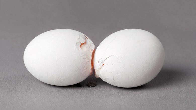 Two white broken eggs together as a little bit of the egg white drips out against a gray background.
