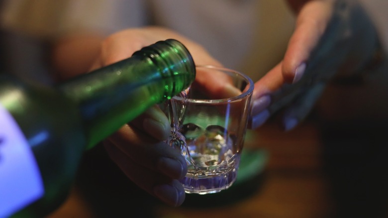 A bottle pouring soju into a shot glass held by hands