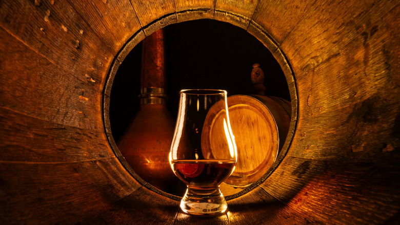 A glass of Scotch whisky sitting inside a wooden barrel, with another barrel and a still visible in the background