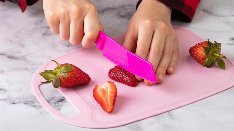 A child cuts strawberries with a pink plastic knife on a pink plastic cutting board