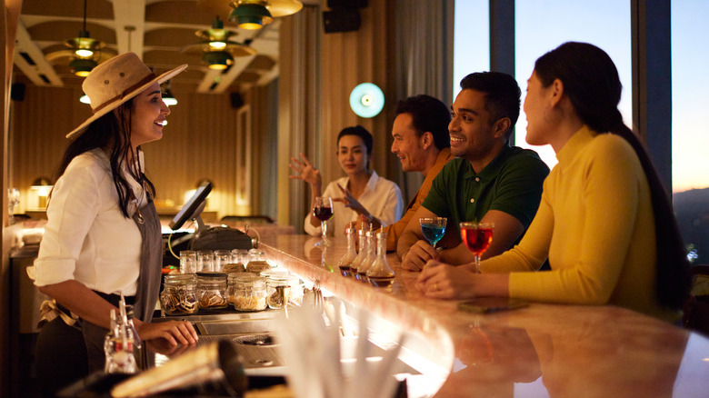 A bartender wearing a hat talking to guests drinking colorful drinks sat at the bar.
