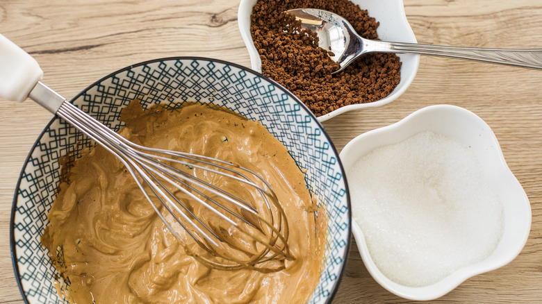 A bowl of whipped coffee with a whisk inside next to bowls of coffee granules and sugar