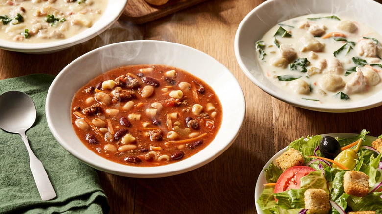 White bowls of various soups from Olive Garden next to bowl of salad on wooden table