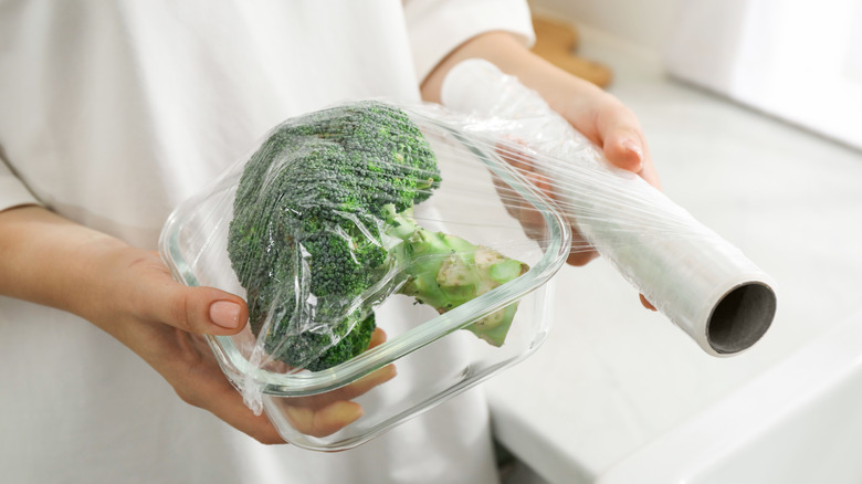 Woman rolling plastic wrap over broccoli in container