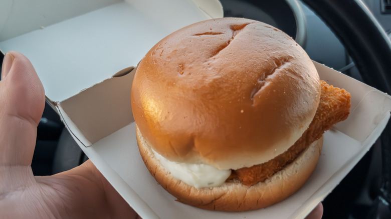 Hand holding a Filet-O-Fish sandwich in its open box in front of a car's steering wheel