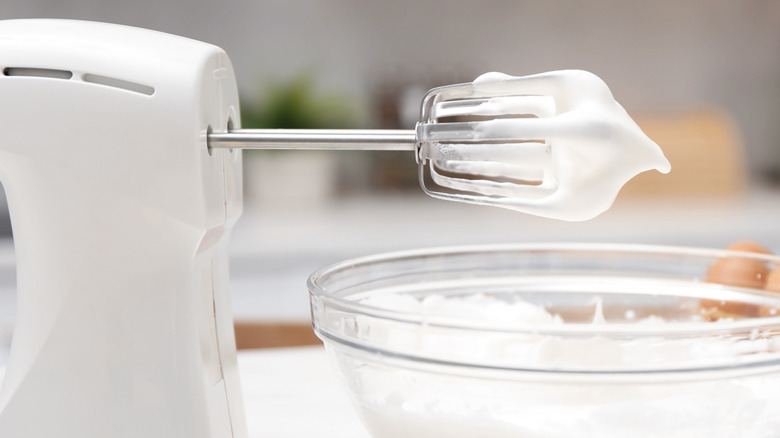 An electric hand whisk with whipped cream on it over a bowl of cream