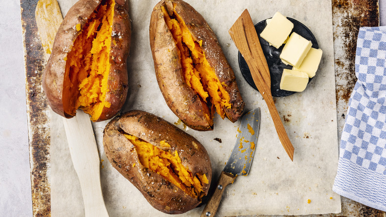 Three baked sweet potatoes on a baking sheet with butter and salt