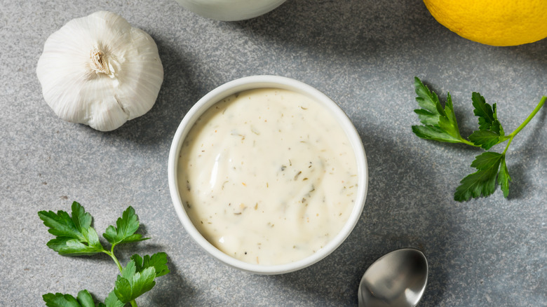 A bowl of homemade ranch dressing on a table next to a spoon, parsley, garlic, and lemon