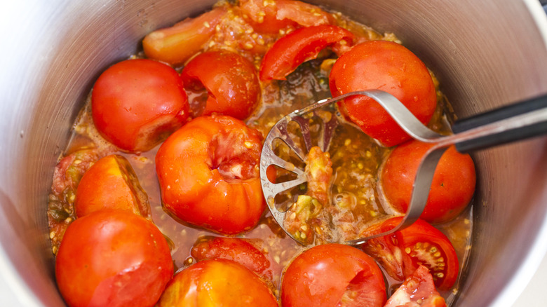 A person mashing up cooked tomatoes using a metal potato masher tool
