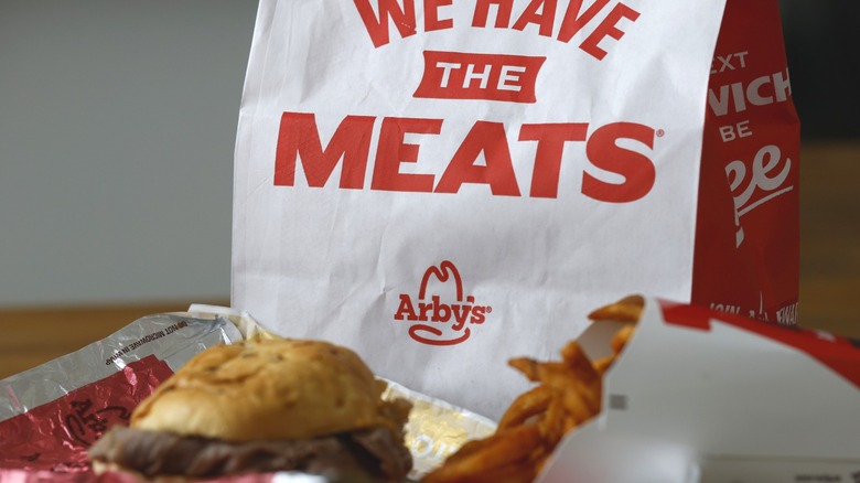 A bag of Arby's food with a sandwich and fries in the foreground sitting on a takeout tray