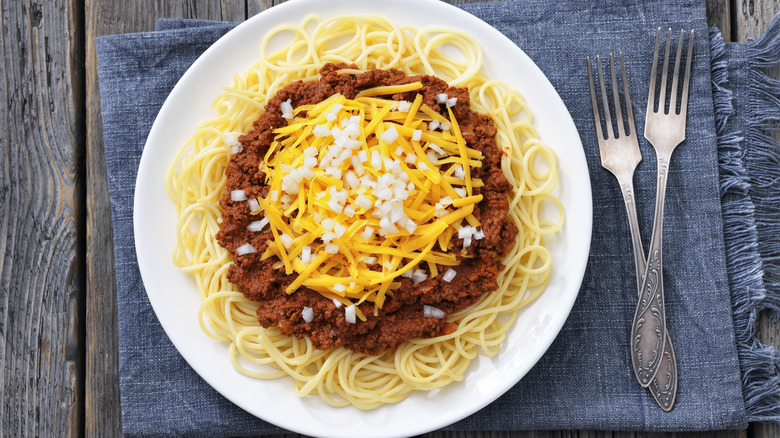 A plate of Cincinnati chili on a mat on a wooden table, garnished with onion, with two forks beside it