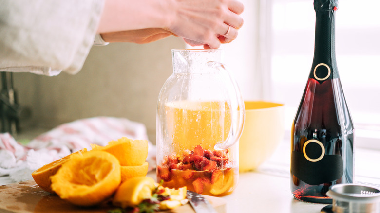 Hands squeeze an orange into a jug of fruit next to a a bottle of prosecco and squeezed oranges.