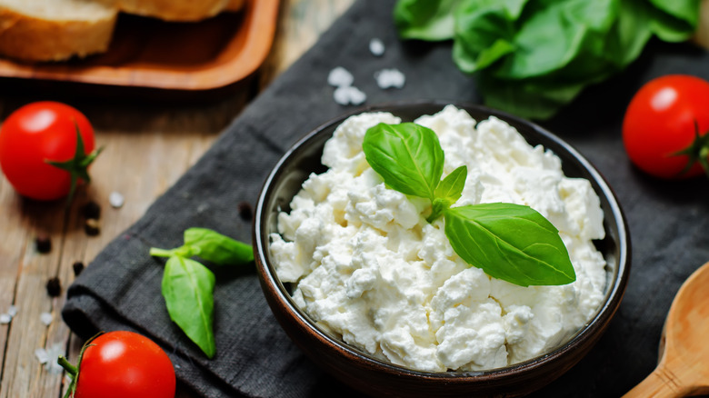 A bowl of ricotta cheese with a basil leaf on a black linen napkin with cherry tomatoes and other items surrounding it.
