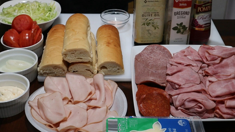 The ingredients for Italian subs laid out on a table, including submarine rolls, cold cuts, condiments, lettuce, and tomatoes