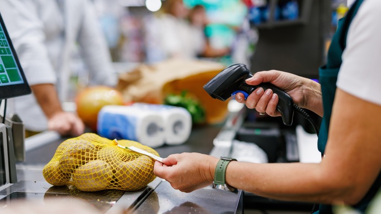 Supermarket cashier scanning potatoes at the checkout using a barcode scanner, with a customer waiting and other shoppers in the background