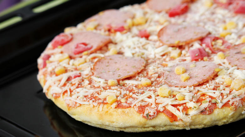 Frozen pizza with salami and corn on baking sheet.
