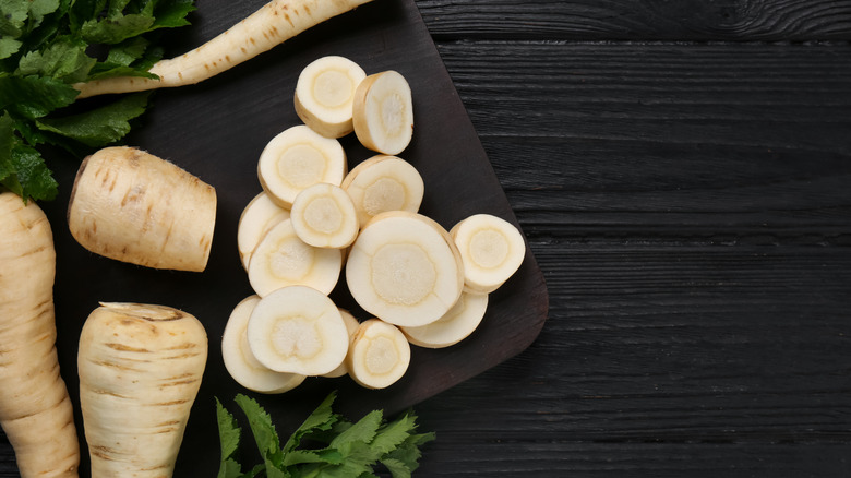 Sliced parsnip discs on a black cutting board with whole parsnips nearby.