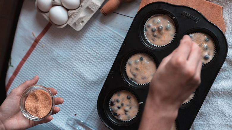 Hands sprinkling coarse sugar over a pan of blueberry muffin batter