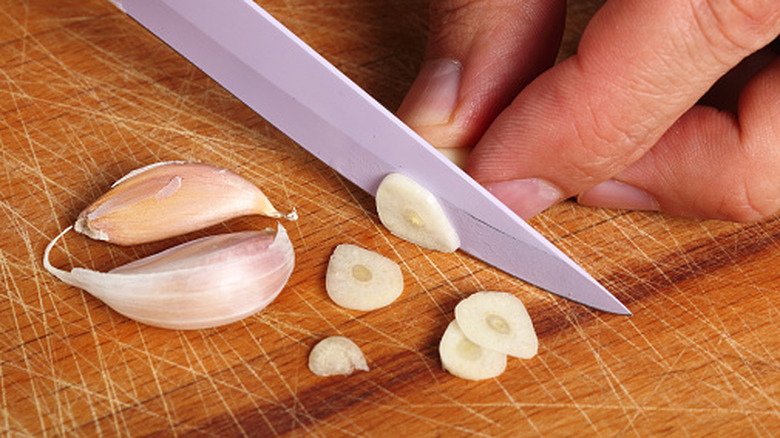 One hand holds a clove of raw garlic in place on a chopping board while a blade thinly slices the garlic