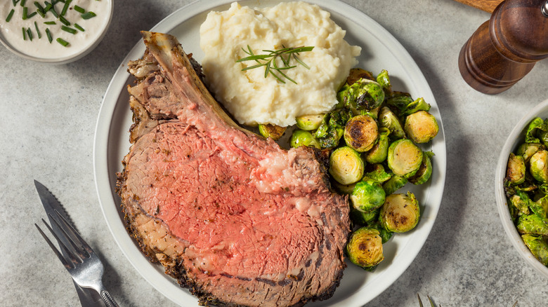 A large slice of prime rib on a white plate, along with mashed potatoes and roasted Brussels sprouts, beside a knife and form, a pepper grinder, and some side dishes