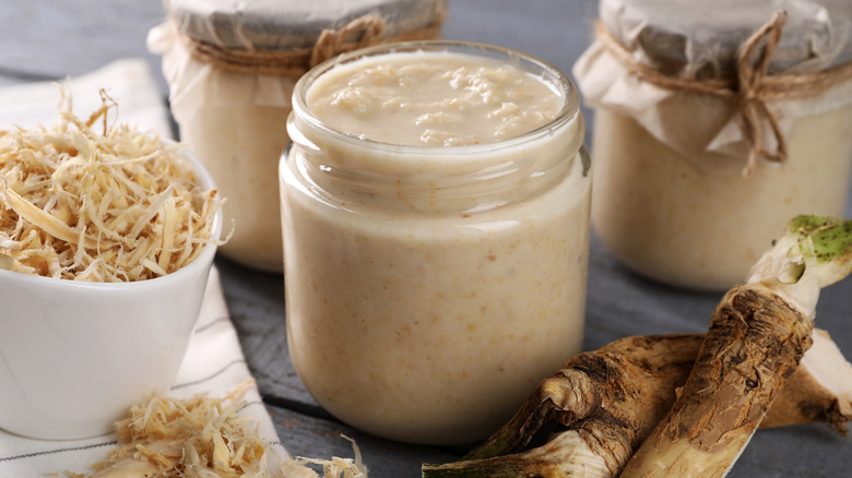Jar of prepared horseradish next to grated and fresh horseradish on black surface