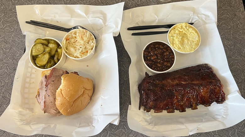 Two paper-lined trays with brisket sandwich, short ribs, and barbecue sides on restaurant table