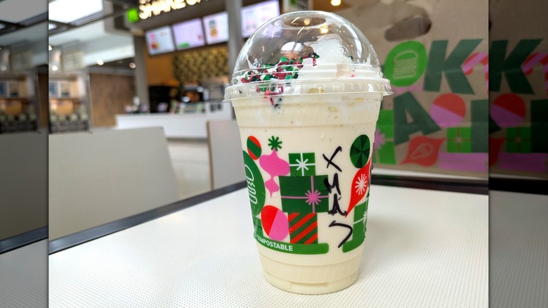 A disposable cup is filled with a white milkshake topped with red and green sprinkles and whipped cream