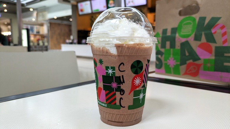A dark brown chocolate milkshake is in a disposable cup with whipped cream on top