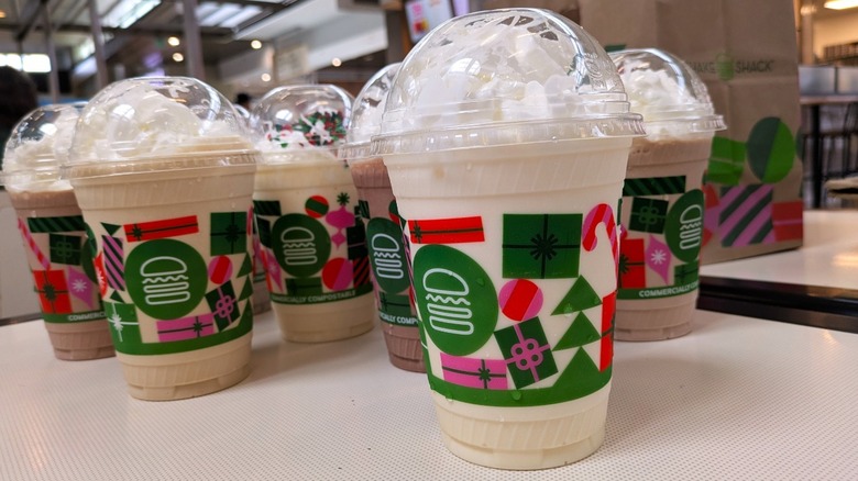 Several milkshakes in holiday-themed disposable cups from Shake Shack are arranged on a table with the Shake Shack sign slightly visible in the background