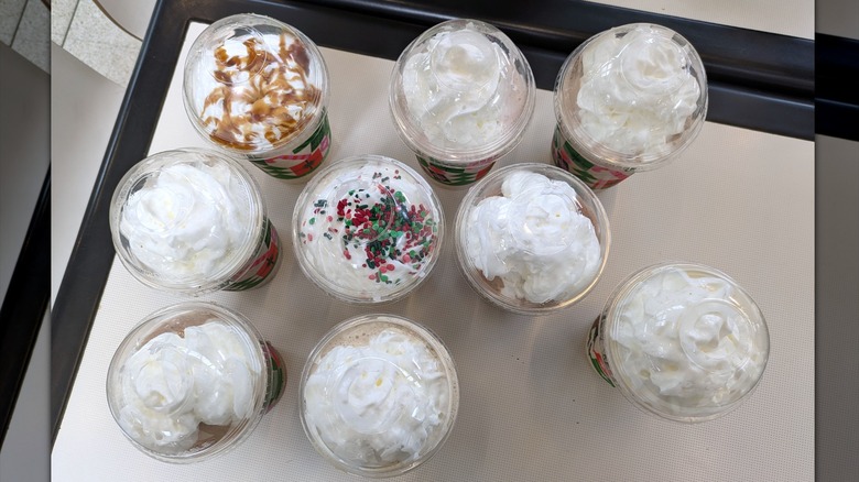 Nine milkshakes are seen arranged on a table from a bird's eye view. There is whipped cream on top of all nine, but only two have additional toppings. One is drizzled with a caramel brown toffee sauce and the other is heavily sprinkled with red and green colored sprinkles.