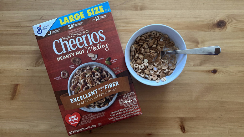 Top view of Cheerios Maple Cinnamon Hearty Nut Medley cereal in a white bowl with milk with a box next to it on a table