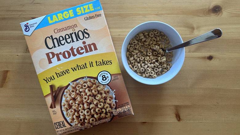 A box of Cheerios Cinnamon Protein cereal box with a white bowl on table filled with cereals