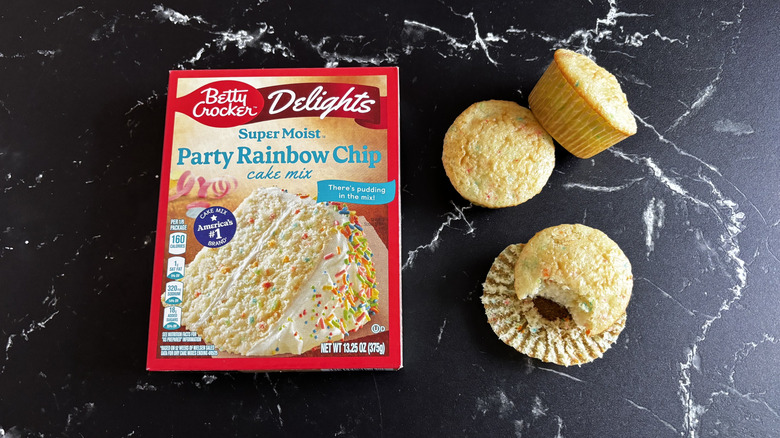 Betty Crocker party rainbow chip cake mix box next to cupcakes made with the mix laid on a black marble counter