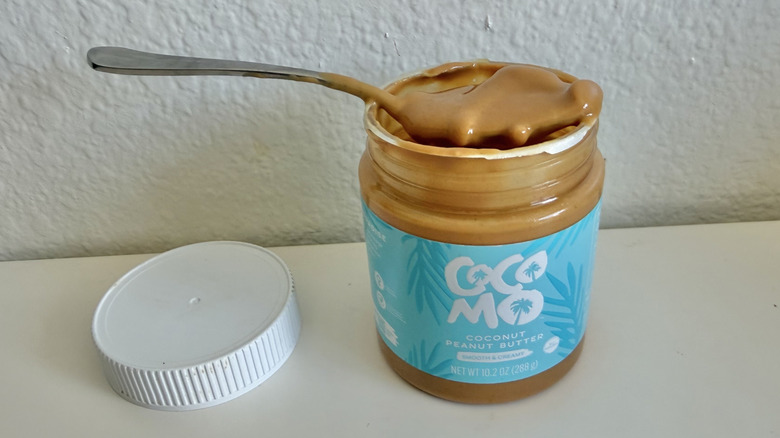 Cocomo Coconut peanut butter opened on a white table with a spoon filled with peanut butter on top of the jar