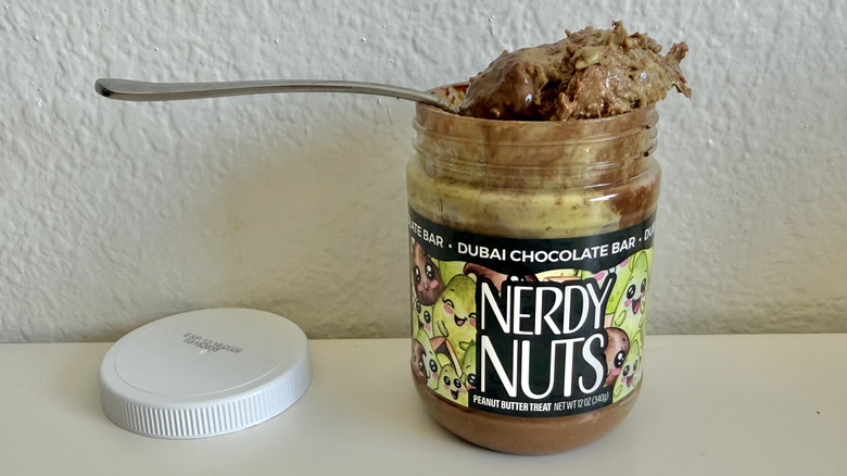 Nerdy Nuts Dubai Chocolate peanut butter jar with a spoon nestled on top filled with peanut butter