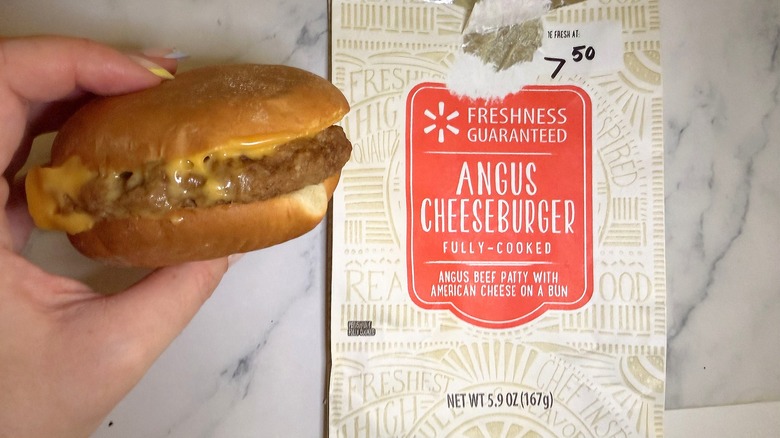 Hand holding Walmart deli cheeseburger next to wrapper on marble surface