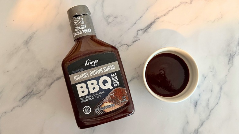 Bottle of Kroger Hickory Brown Sugar BBQ Sauce on table next to small white bowl of sauce