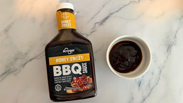 Bottle of Kroger Honey Sweet BBQ Sauce on table next to small white bowl of sauce