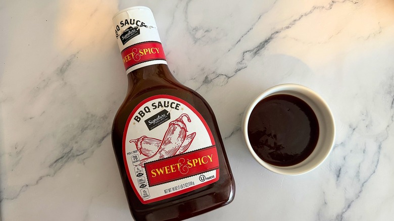 Bottle of Signature Select Sweet & Spicy BBQ Sauce on table next to small white bowl of sauce
