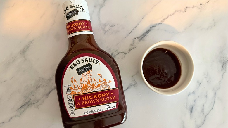 Bottle of Signature Select Hickory & Brown Sugar BBQ Sauce on table next to small white bowl of sauce