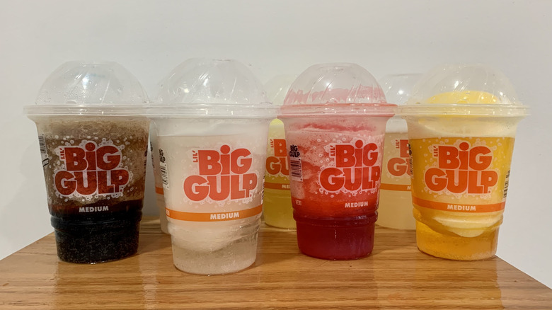 eight different Slurpees in plastic cups on wooden shelf with white background