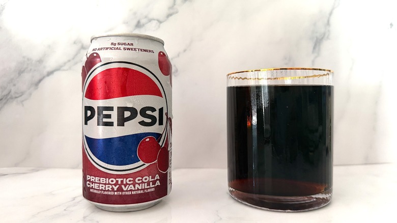 Can of Pepsi Prebiotic Cola Cherry Vanilla next to gold-rimmed glass of Pepsi on marble surface