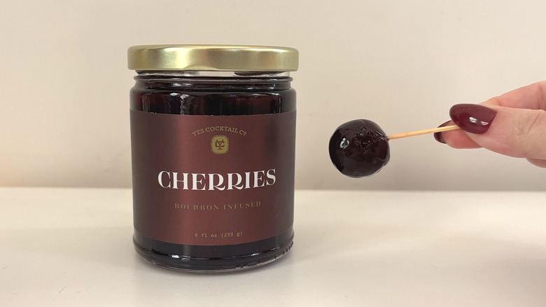Jar of Yes City Cocktail Co. cocktail cherries next to hand holding cherry on toothpick
