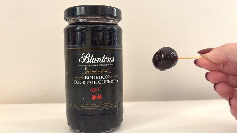Jar of Blanton's bourbon cocktail cherries next to hand holding cherry on toothpick