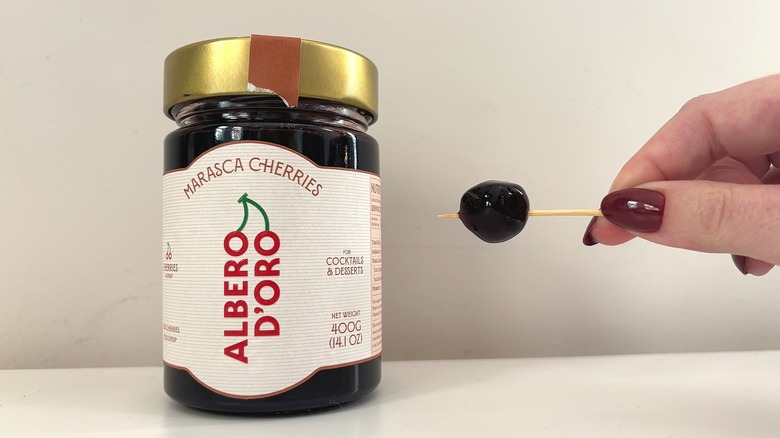 Jar of Albero D'Oro cocktail cherries next to hand holding cherry on toothpick