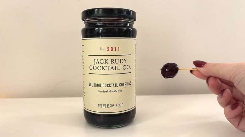 Jar of Jack Rudy Cocktail Co. Bourbon cocktail cherries next to hand holding cherry on toothpick