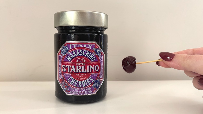 Jar of Hotel Starlino cocktail cherries next to hand holding cherry on toothpick